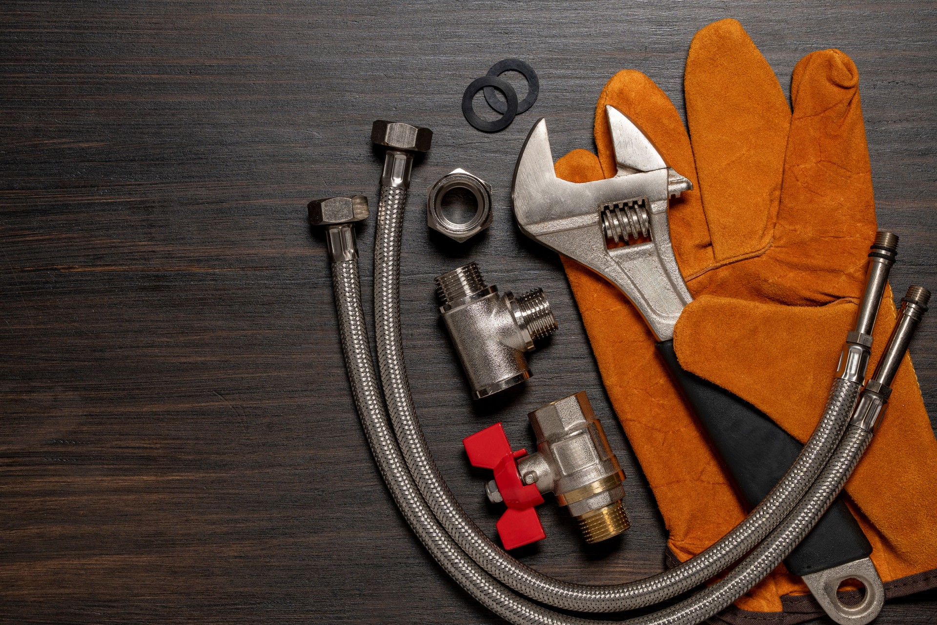 Adjustable wrench, ball valve with red handle, stainless steel tee fittings, leather safety gloves and rubber gaskets on wooden background, top view, copy space, closeup. Plumbing repair concept